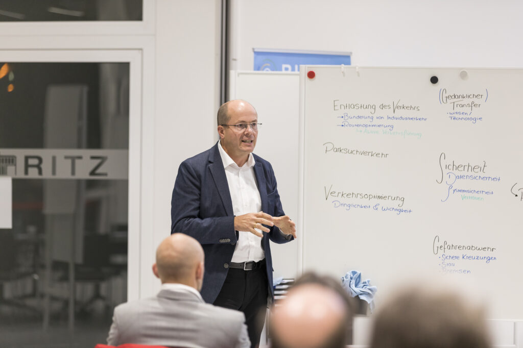 Dr. Walter Naumann (ETO) hält im RITZ einen Vortrag vor Publikum. Im Hintergrund steht eine Whiteboard-Wand mit handschriftlich notierten Themen wie „Verkehrsoptimierung“, „Sicherheit“ und „Gedanklicher Transfer“.