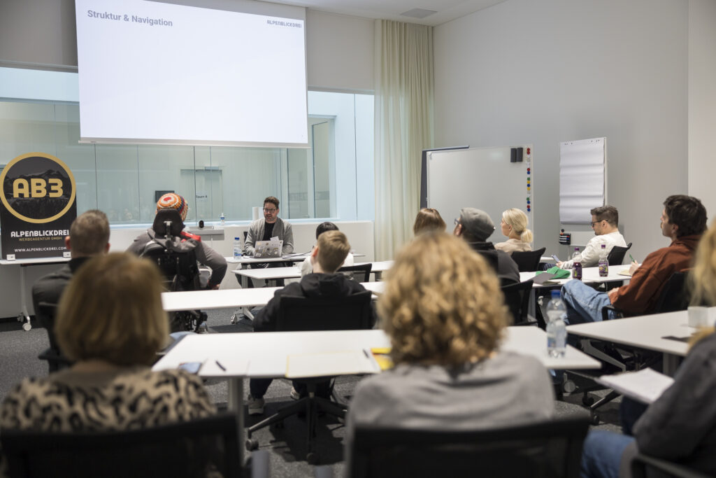 Vortrag im RITZ Friedrichshafen: Ein Referent der Agentur „Alpenblickdrei“ spricht vor einem aufmerksam zuhörenden Publikum über Struktur und Navigation hinsichtlich digitaler Barrierefreiheit. Auf der Leinwand ist eine Präsentationsfolie zu sehen.