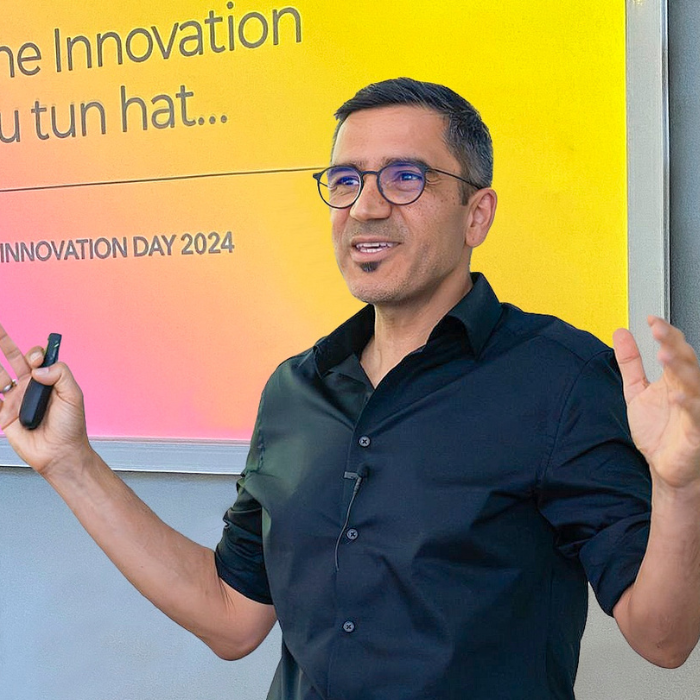 Hansjoerg Probst spricht vor einem leuchtend gelben bis pinken Hintergrund mit der Aufschrift „INNOVATION DAY 2024“. Er trägt ein schwarzes Hemd, Brille und hält einen Presenter in der Hand.