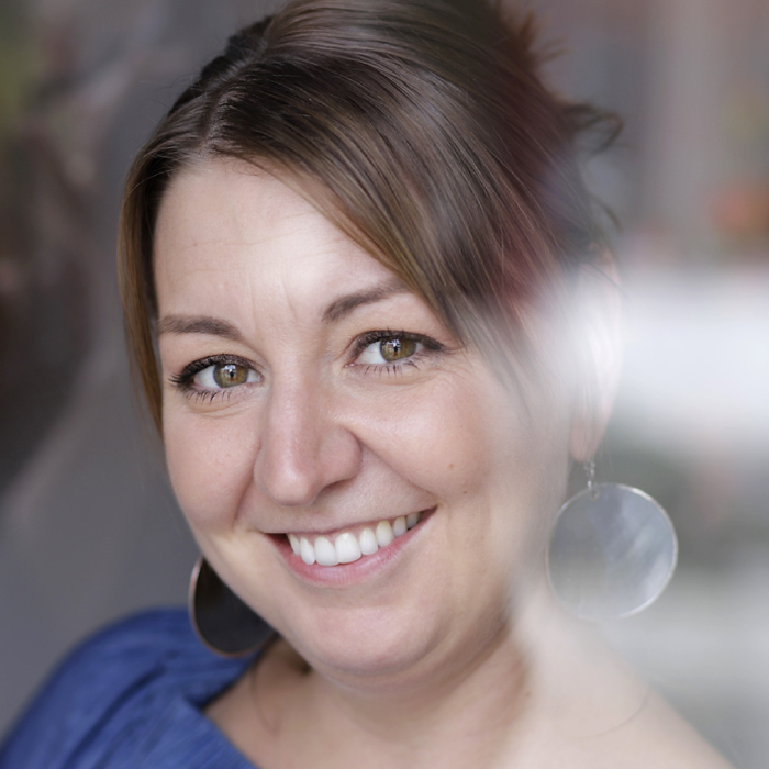 Porträt von Julia Kröll, lächelnd, mit braunen Haaren und großen runden Ohrringen, vor unscharfem Hintergrund.