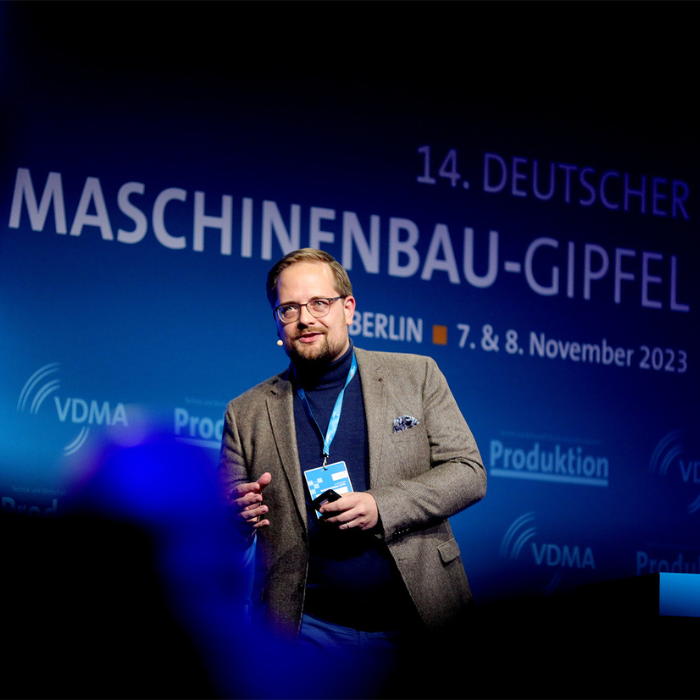 Dr. Ulrich Hutschek spricht auf der Bühne des 14. Deutschen Maschinenbau-Gipfels in Berlin am 7. und 8. November 2023. Er trägt ein Headset-Mikrofon und hält eine Präsentationsfernbedienung in der Hand. Im Hintergrund ist das Veranstaltungsbranding mit Logos von VDMA und „Produktion“ sichtbar.