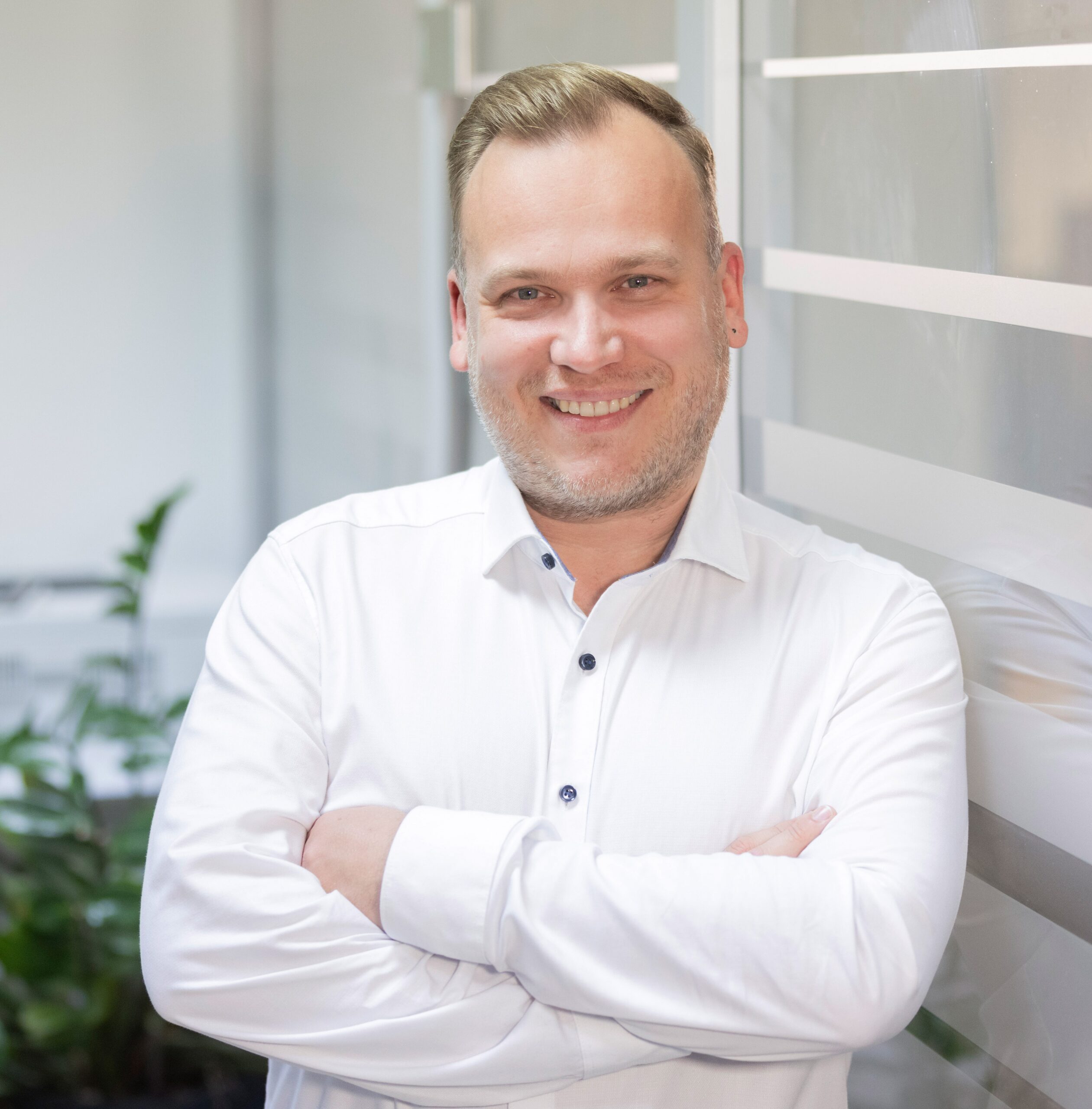 Matthias Ruf, Geschäftsführer der all for IT GmbH, steht lächelnd mit verschränkten Armen in einem modernen Büroflur. Er trägt ein weißes Hemd, im Hintergrund sind Glaswände und grüne Pflanzen zu sehen.