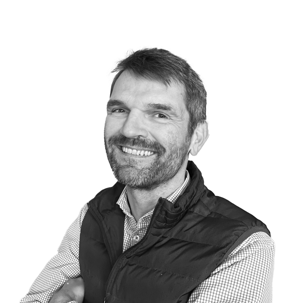 Schwarz-weiß-Porträt von Dr. Mario Herger. Er lächelt in die Kamera, trägt ein kariertes Hemd und eine dunkle Steppweste. Der Hintergrund ist neutral und strukturiert.