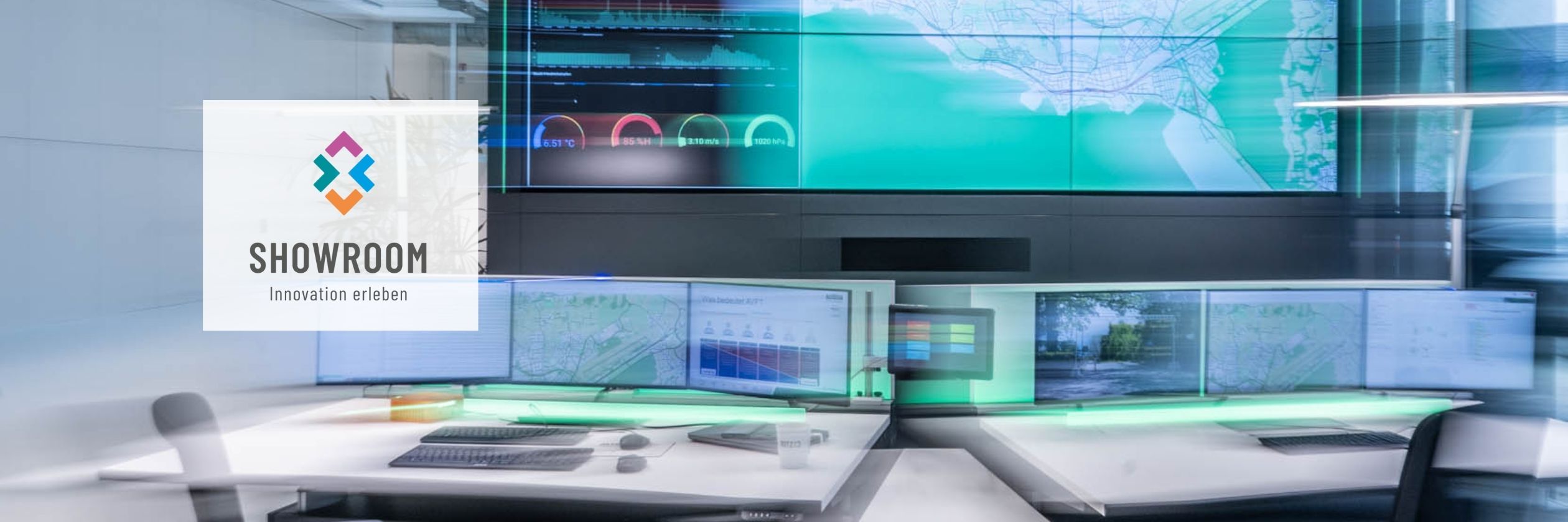 Zwei moderne Arbeitsplätze mit jeweils mehreren Monitoren und Tastaturen, angeordnet vor einer großformatigen Videowand. Die Videowand zeigt Karten, Diagramme und grafische Benutzeroberflächen in überwiegend grünen Farbtönen. Der Raum ist dunkel gehalten und wird durch das Licht der Bildschirme sowie eine grün leuchtende Lichtleiste unter den Monitoren beleuchtet. Links im Vordergrund ist ein Stuhl angeschnitten.