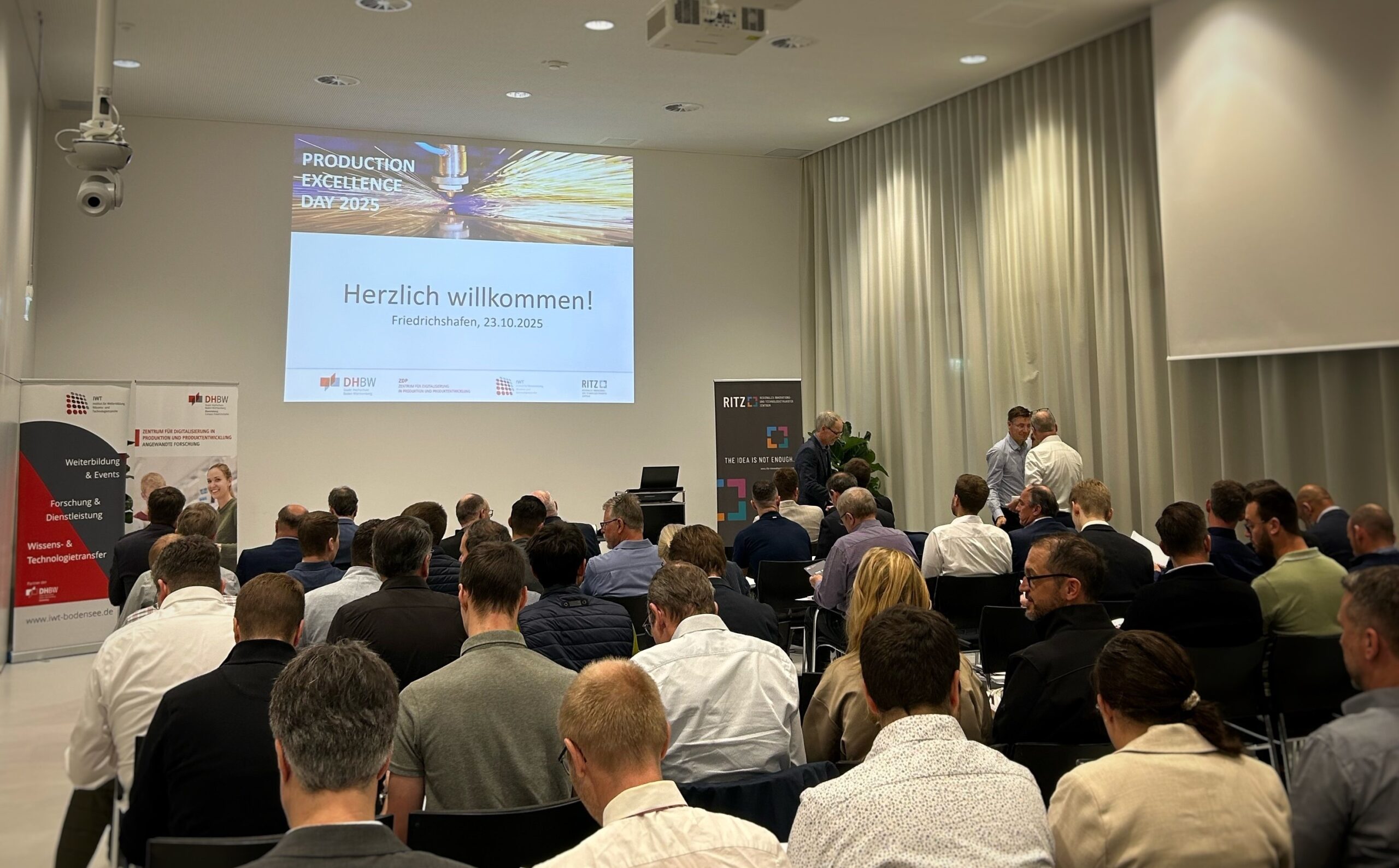 Teilnehmerinnen und Teilnehmer sitzen in einem gut gefüllten Veranstaltungssaal und warten auf den Beginn des Production Excellence Day 2025. Auf der Leinwand vorne steht ‚Herzlich willkommen! Friedrichshafen, 23.10.2025‘. Links und rechts stehen Roll-ups der Veranstalter, darunter DHBW, IWT und RITZ.