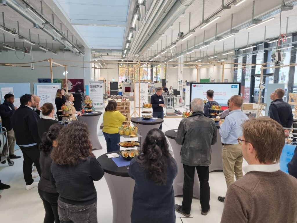Netzwerktreffen mit Präsentation in einer modernen Lern- und Produktionshalle, Teilnehmende stehen an Stehtischen und verfolgen einen Fachvortrag.