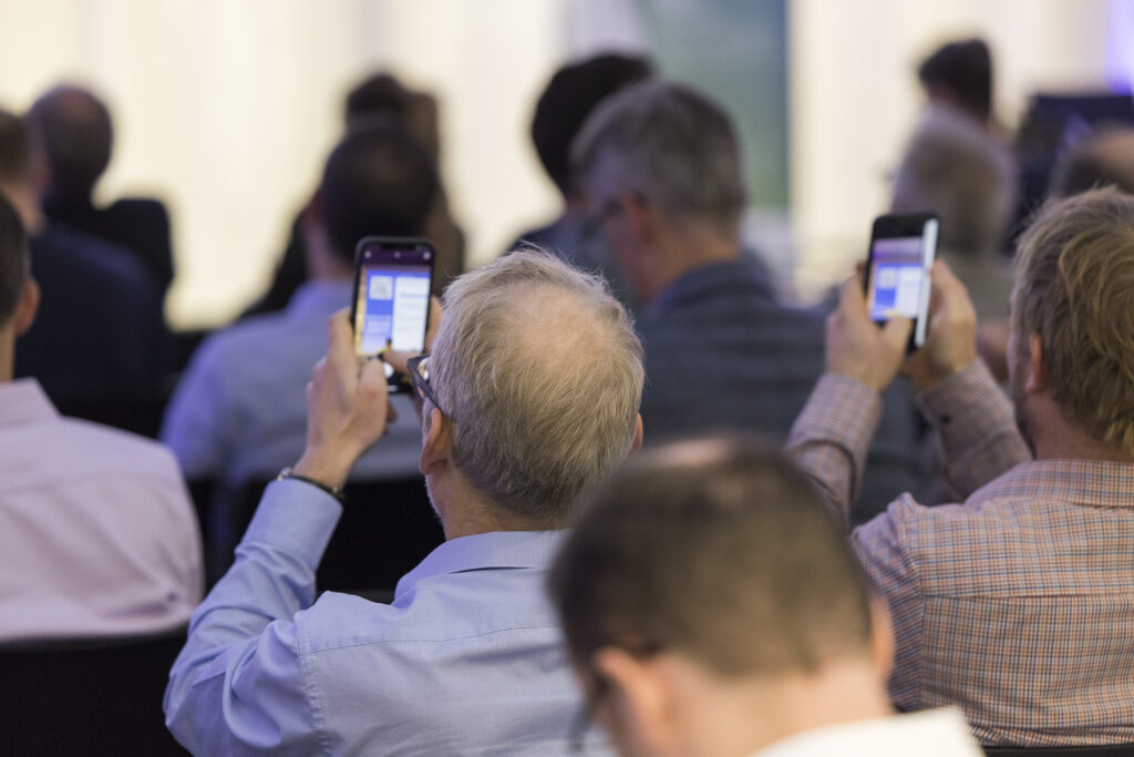 Teilnehmende halten Smartphones hoch und beteiligen sich digital an einer Interaktion bei der Veranstaltung DRIVING CHANGE im RITZ.