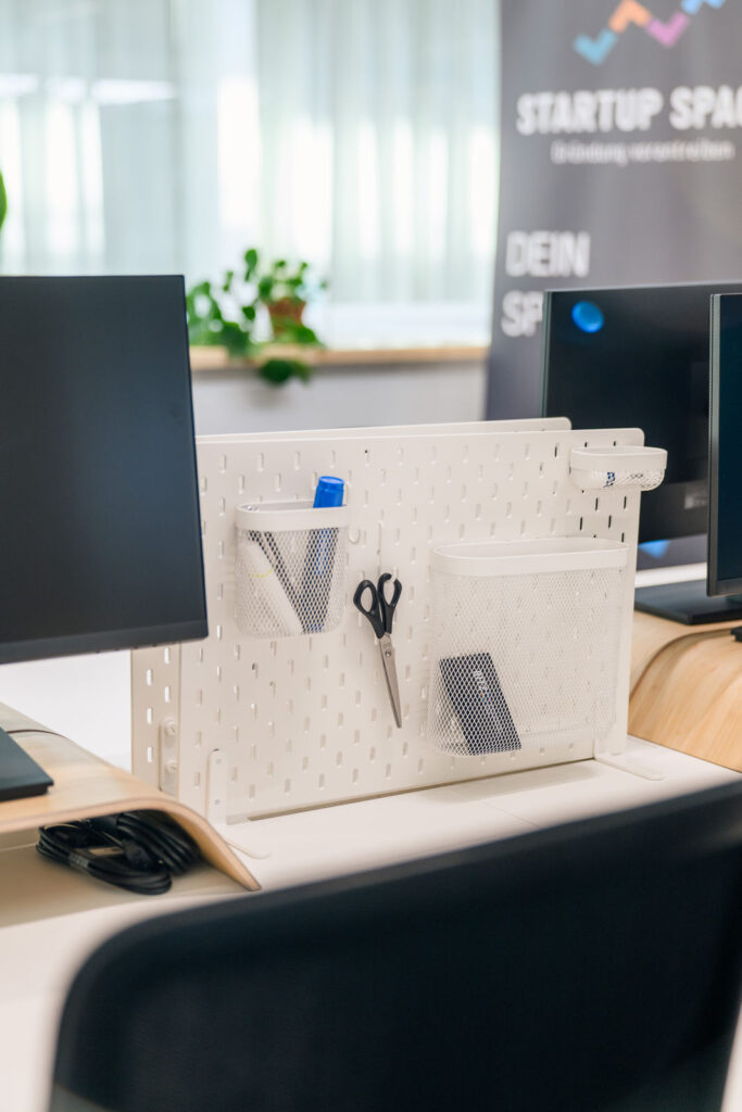 Nahaufnahme Arbeitsplatz im Startup Space mit Organizer-Pinnwand, Monitoren und Roll-up „Startup Space“